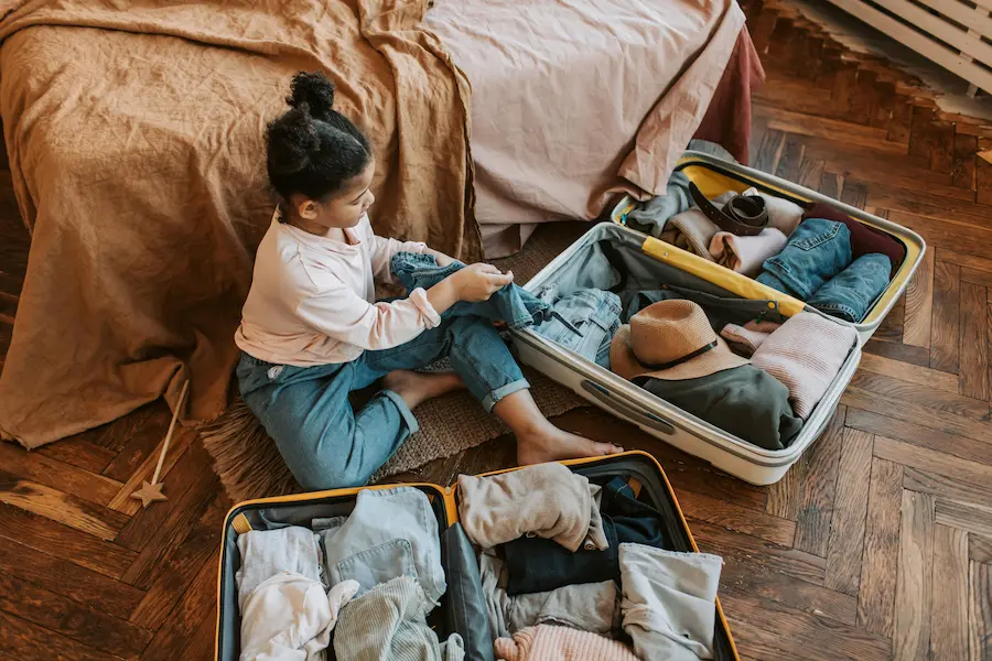 A toddler and preschooler are sitting on the floor surrounded by open suitcases and packing cubes for a trip to Asia.